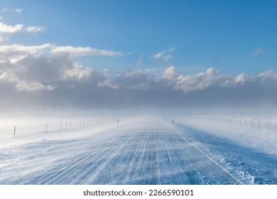 View over snow covered road in agricultural landscape in Iceland with high winds blowing snow over road creating a blur with snow clouds in distance giving way to clear blue sunny sky