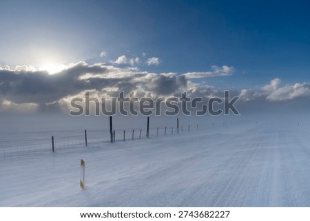 View over snow covered through agricultural snowy landscape with high winds blowing snow over road in Iceland with snow clouds in distance giving way to clear blue sunny sky