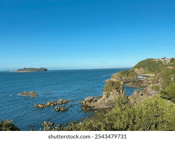 View of the Pacific Ocean from Ancud, Chiloé, Chile, with rocky shoreline and a distant island. The scene shows clear blue skies, calm waters, and lush green hills creating a serene coastal landscape - Powered by Shutterstock