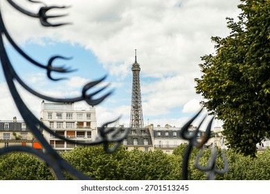 A view of part of the Eiffel Tower, including its top, seen through a fence. Various buildings are in the foreground. The sky is blue and cloudy.