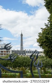 A view of part of the Eiffel Tower, including its top, seen through a fence. Various buildings are in the foreground. The sky is blue and cloudy.