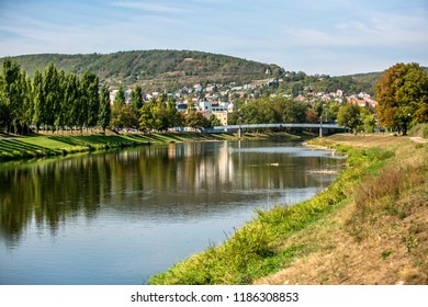 View Of Picturesque Landscape Of Green Hill, Town Beroun, Czech Republic, Europe, Bridge Over River Berounka, Dry Grass River Bank, Reflection Of Trees In Water, Sunny Fall Day, Blue Sky, White Clouds