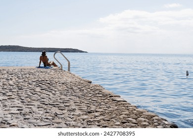 View of pier in the south of the island of Crete with a bather sitting sunbathing nest to the stairs contemplating the sea