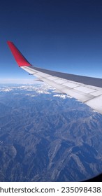 View from the plane window, big wing of the plane, white with red detail over beautiful mountains with remnants of snow, beautiful sunny day with clear blue sky. Clouds on the horizon. Vertical photo.