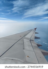 View from a plane window looking out over the large metal wing against a blue sky, white clouds, and distant horizon over the sea.