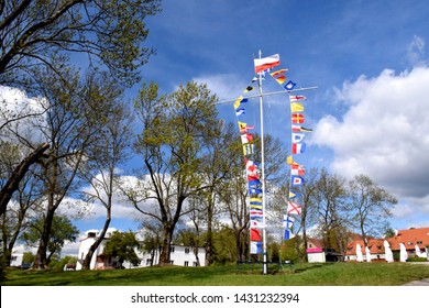 View of a pole with numerous flags, including the one of Poland, France, England, Ireland, and Japan seen during a windy day on a field surrounded by massive trees and some houses during a cloudy day