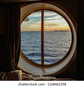 A view from the porthole window of a cruise ship, showing the city buildings in the sunset. The photo was taken inside a cabin on a Caribbean cruise.