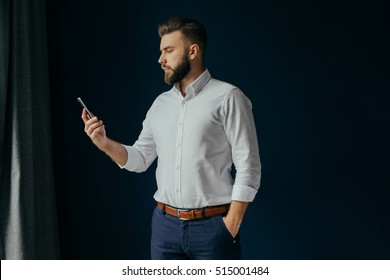 View in profile of a young bearded businessman, dressed in a white shirt, standing in the center of the frame near the window on a dark-blue walls and looking at the screen of smartphone in his hand.