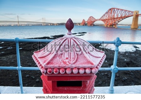 View of a red pillar box covered in frost with the George IV Rail Bridge, Firth of Forth Road bridge and Queensferry Crossing Bridge on the background under a blue sky in a cold Winter day