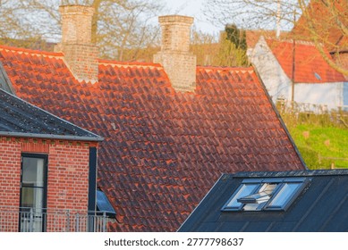 View of red tiled rooftops with brick chimneys and a modern skylight window in a peaceful residential area surrounded by trees and greenery.