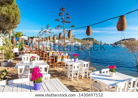 View of restaurant or cafe and bougainvillea flowers on beach in Gumusluk, Bodrum city of Turkey. Aegean seaside style colorful chairs, tables and flowers in Bodrum town near beautiful Aegean Sea.