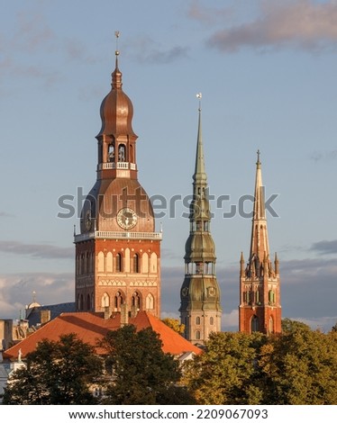A view of Riga with three church towers and trees. The steeples of Riga Cathedral, St. Peter's Church and St. Saviour's Church In Riga Old Town can be seen behind trees.