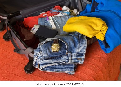 View Of A Rigid Suitcase With A Variety Of Brightly Colored Clothes Half Placed Inside With Accessories Such As A Watch And A Test For The Covid-19. Holiday Travel Concept. Summer Holidays.