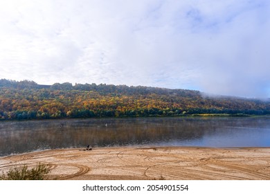 A view of the river between two banks, a fisherman on the shore and in a boat, a colorful forest on the opposite shore. Autumn river landscape. Morning fog over the water, clouds