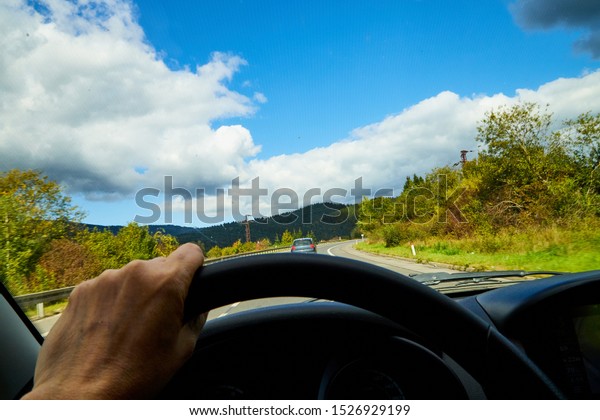 View of the road with a beautiful mountain landscape\
from the car window in a nice summer or autumn day. Woman\'s hand on\
the steering wheel. Female driver and beautiful landscape during\
travel in auto
