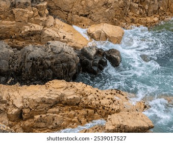 View of rocky coastline with waves crashing against the rocks. The scene features textured rocks in various shades of brown and gray, surrounded by clear turquoise water. - Powered by Shutterstock