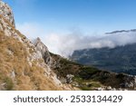 View of a rocky mountain slope on a clear day, with golden grassy terrain leading towards cloud-covered valleys below. Serene landscapes, mountain exploration, and tranquility in the wilderness.