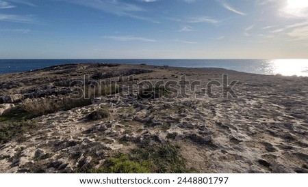 view of the rocky shore and town from nordsee in malta, sea on left side, buildings at right side, sky is blue with few clouds, camera angle looks over rocks to city, daylight, photo taken by phone