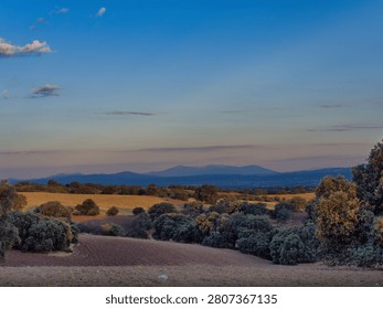 A view of rolling fields and hills at sunset shows trees scattered across the landscape. Mountains are visible far off in the distance. The sky changes colors as the sun sets.