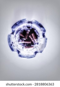 ​Top view of a round blue-patterned ceramic ashtray filled with burnt cigarette butts and ash isolated on a clean white background