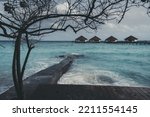 View of a row of resort bungalows with triangle roofs in the background; a tropical tree over a concrete pier and a groyne, a warm teal ocean water of a luxury spa in the Maldives in the foreground