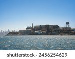 View of rugged Alcatraz Island in San Francisco Bay, California, USA. Shows main cellhouse, guard tower, choppy waters, and city skyline in background.