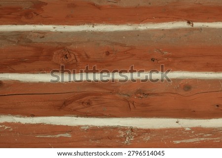 Close‑up view of a rustic log wall showing horizontal wooden beams with visible knots and grain patterns. Light‑colored chinking fills the gaps between the reddish‑brown logs, highlighting the craftsmanship of traditional cabin construction. The texture and natural imperfections of the wood evoke warmth and heritage in rural architecture.