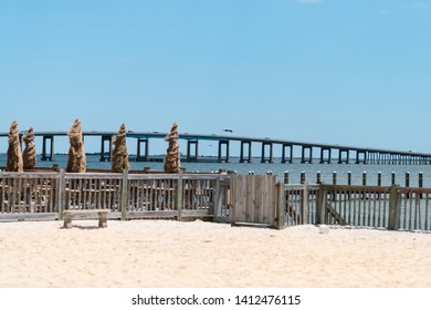 View From Sand Beach In Navarre On Pensacola Bay Bridge With Restaurant Wharf, Fence And Straw Umbrellas In Gulf Of Mexico, Florida Panhandle