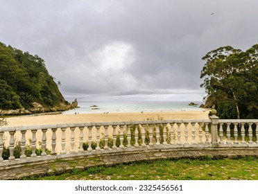 View of a sandy beach with rock cliffs from over a stone balcony covered in grass.