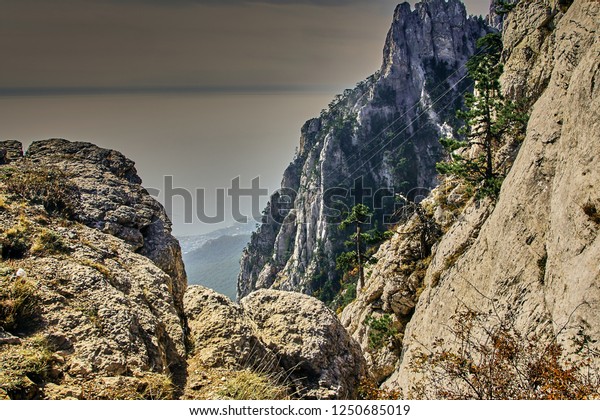 View of the sea through the rocks from the\
highest mountain of Crimea (Ai-Petri mountains). Evergreen pines\
grow on the rocks. Sea in a haze. The sky with white clouds.\
Crimean mountain landscape