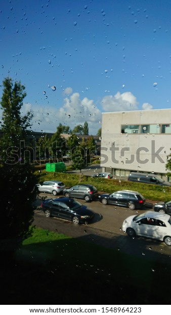 View from the second floor through glass window\
with close-up water droplets on it, street is outside with cars,\
plants, buildings and blue\
sky.