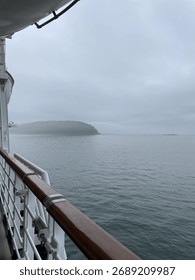 View from a ship deck over calm foggy waters toward a distant island, with muted tones and sky above.