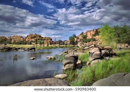 A view from the shore of Watson Lake with blue cloud filled sky and green trees with rock cliffs in Prescott, Arizona.