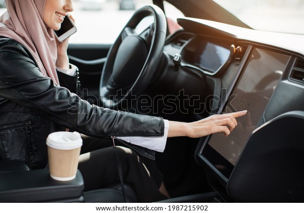 View from shoulder to arabian woman in hijab\
sitting on driver\'s seat of luxury electric car and looking for\
direction on electronic dashboard. Concept of people, vehicle and\
modern technology.