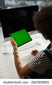 View From The Shoulder Of Woman Holding And Using Hand Gestures Swipe Up On Green Mock-up Screen Digital Tablet Computer Sitting At The Desk. Browsing The Internet. Background Office.