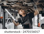 View from side of concentrated worker of car service checking running gear of automobile. Male mechanic in uniform and gloves standing under car, keeping lamp and looking for breakage