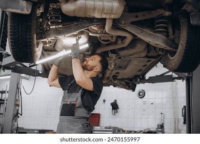 View from side of concentrated worker of car service checking running gear of automobile. Male mechanic in uniform and gloves standing under car, keeping lamp and looking for breakage - Powered by Shutterstock