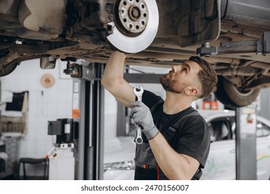 View from side of concentrated worker of car service checking running gear of automobile. Male mechanic in uniform and gloves standing under car and looking for breakage. - Powered by Shutterstock