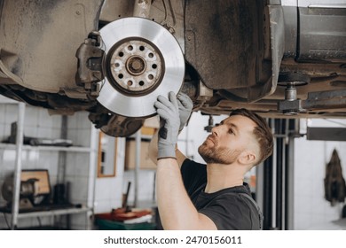 View from side of concentrated worker of car service checking running gear of automobile. Male mechanic in uniform and gloves standing under car and looking for breakage. - Powered by Shutterstock