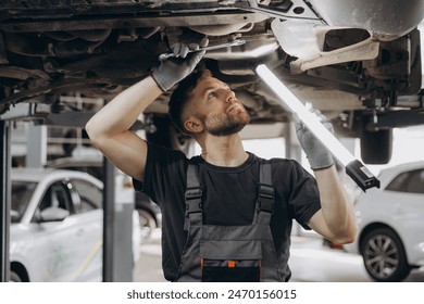 View from side of concentrated worker of car service checking running gear of automobile. Male mechanic in uniform and gloves standing under car, keeping lamp and looking for breakage - Powered by Shutterstock