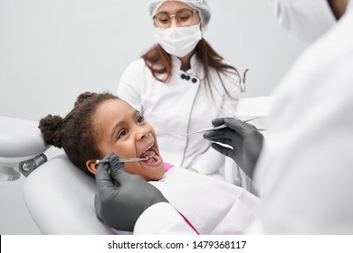 View From Side Of Funny Girl Lying On Dentist Chair With Open Mouth While Doctor Examining Teeth. Pretty African Child Looking At Dentist And Curing Teeth. Concept Of Treatment And Health.