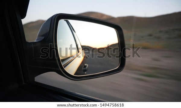 View in the side mirror of the car.\
Orange dawn beyond the hills. The car goes at speed. Visible green\
fields, grass, grasslands. Black color of the\
car.