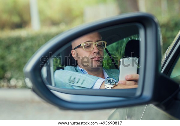 view side mirror reflection of serious young man\
driver looking at phone, texting in car.  Safe trip journey driving\
concept