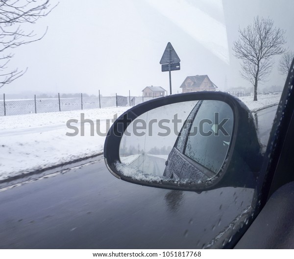 view of the side mirror
while driving from the driver's perspective. Snow on the mirror,
drops of water on the glass. In the background a winter landscape
and snow.