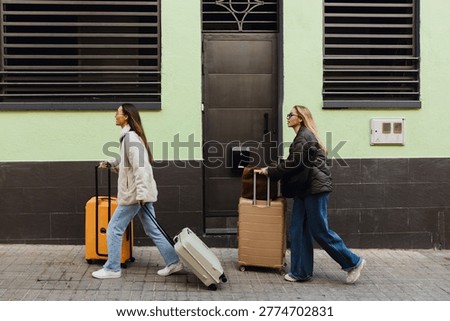 A view from the side of two young White female friends, a blond, and a brunette, walking up an incline on a city street, wheeling their suitcases along, on a sunny day