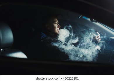 View from the side of a young man smoking an e-cigarette as he drives his car on an urban street. At night, or indoors in the parking lot, backlight