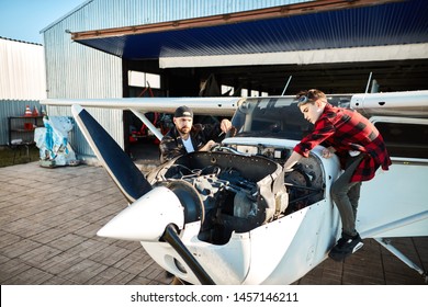 view of small white propeller air jet standing outside hangar building, male pilot with screwdriver stands besides, checking engine and his son helping him with inspecting airplane's systems. - Powered by Shutterstock