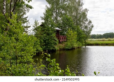 View Of A Small Wooden Cottage By The Lake. The Building Is Also Used By Anglers. Outdoor Recreation After A Working Week, Hobby.