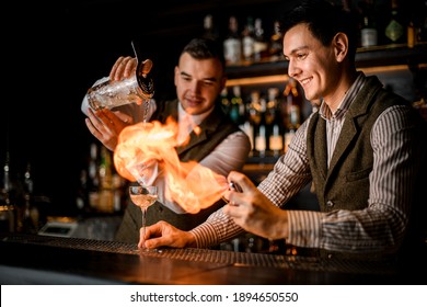 View of smiling bartender man who sets on fire cocktail glass at bar when another barman pours drink into it