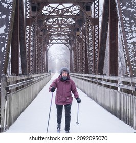 View Of Smiling Older Woman Skiing Cross-country Under Old Railroad Bridge Transferred To State Recreation Trail On Snowy Winter Day In Midwest